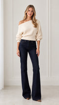 Full front view of model wearing cream asymmetrical one-shoulder ribbed knit sweater with dark denim flare jeans
