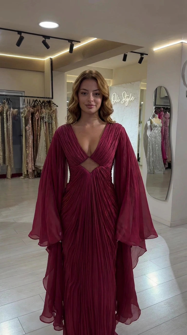 Front view of woman wearing burgundy pleated maxi gown with cutout and long cape sleeves