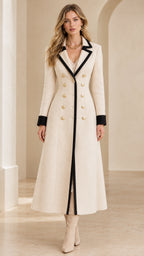 Front full-length view of woman wearing cream maxi coat with black contrast trim and gold crest buttons