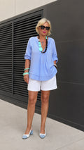 Full front view of model wearing Light Blue pleated split neck tunic blouse with white shorts