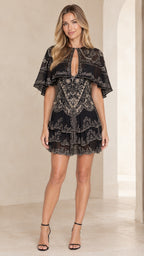 Front full view of woman wearing black embroidered capelet tiered mini dress