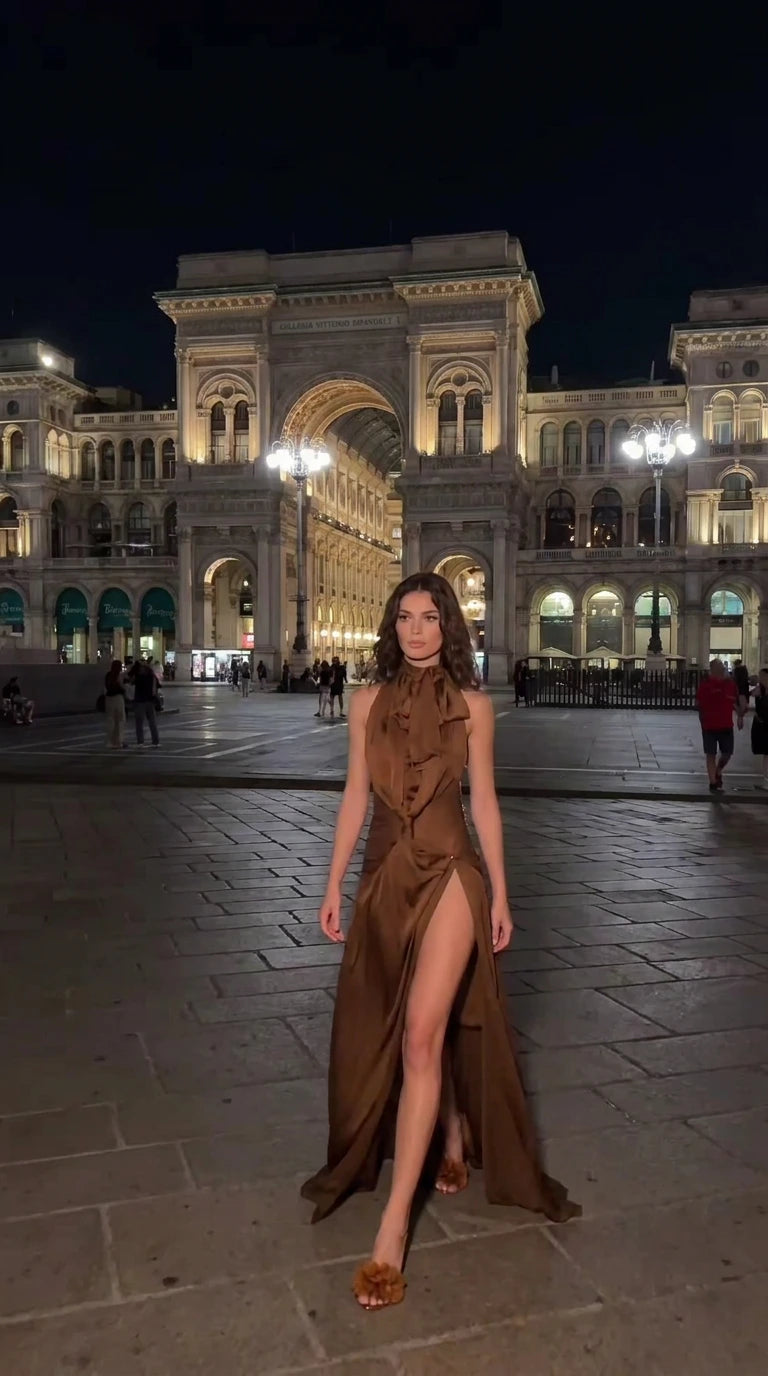 Woman wearing brown satin halter neck maxi dress with ruffled collar and thigh slit in front of historic architecture
