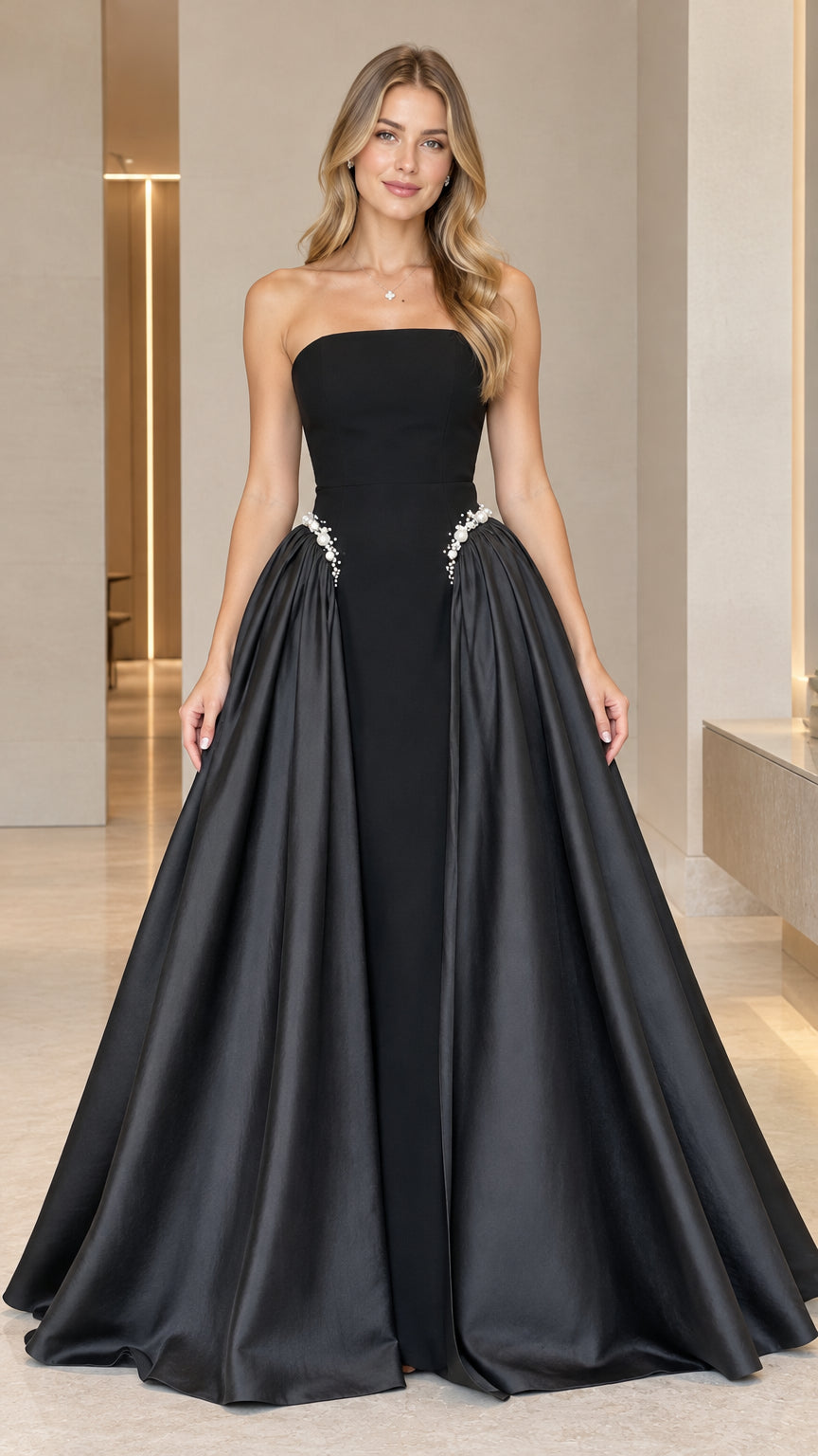 Front full-length view of model wearing black strapless satin evening gown with side overskirt and pearl waist detail