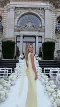 Three-quarter rear view of woman wearing pale yellow corset maxi dress with draped shoulder sash at an outdoor wedding venue