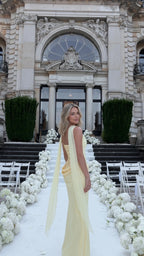 Three-quarter rear view of woman wearing pale yellow corset maxi dress with draped shoulder sash at an outdoor wedding venue