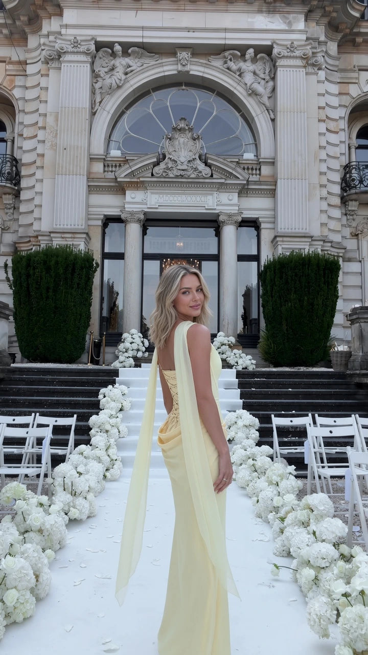 Three-quarter rear view of woman wearing pale yellow corset maxi dress with draped shoulder sash at an outdoor wedding venue