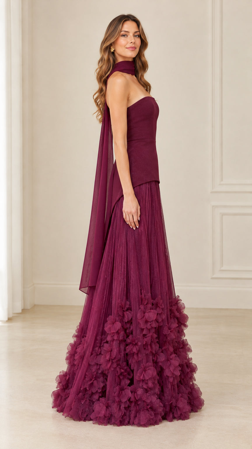 Three-quarter angle of model in burgundy strapless evening gown showcasing floating neck drape and pleated skirt