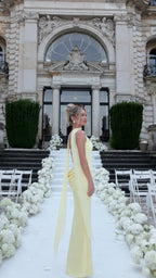 Side profile view of woman wearing pale yellow corset maxi gown showing slim silhouette and trailing shoulder drapery