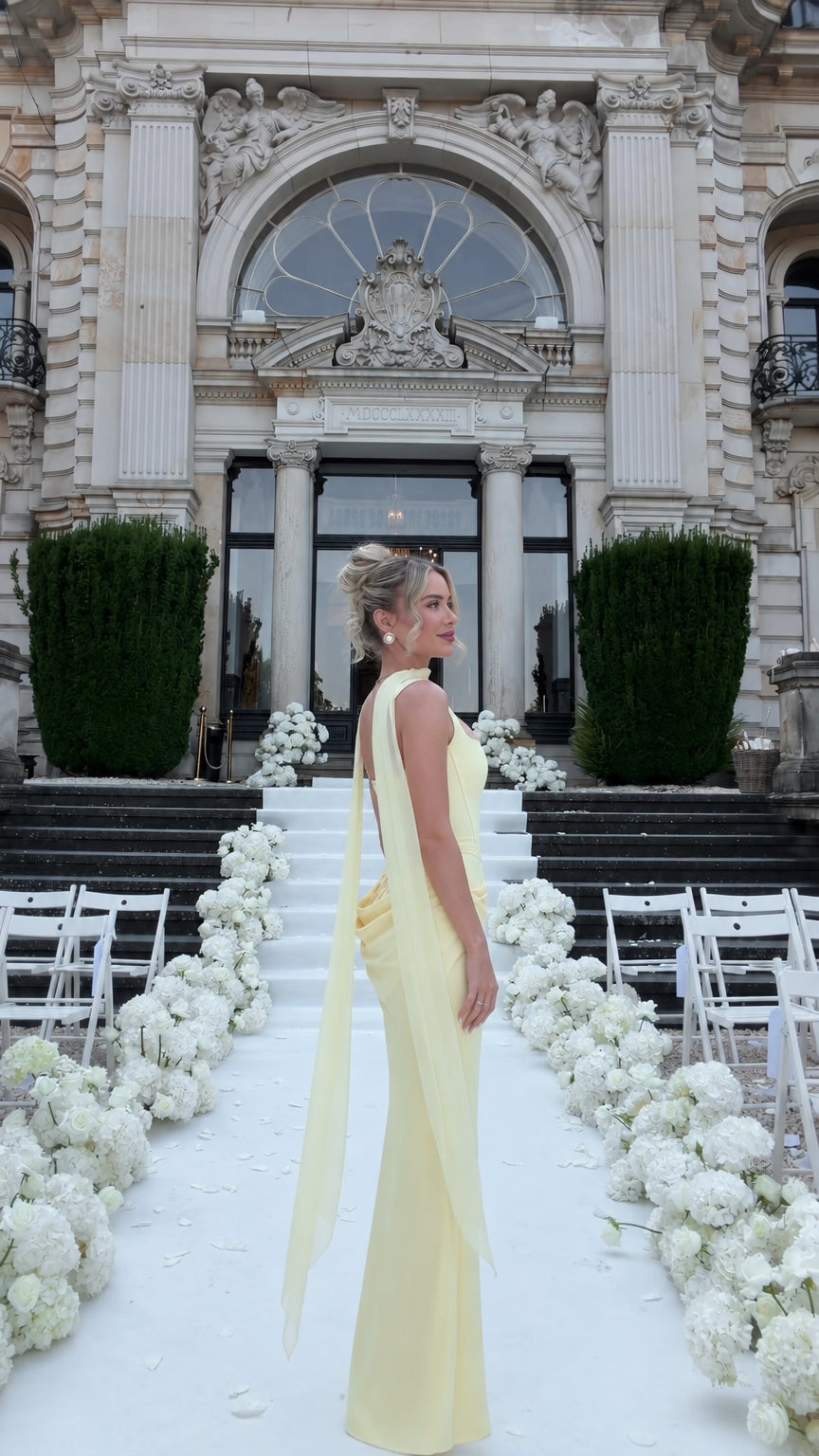 Side profile view of woman wearing pale yellow corset maxi gown showing slim silhouette and trailing shoulder drapery