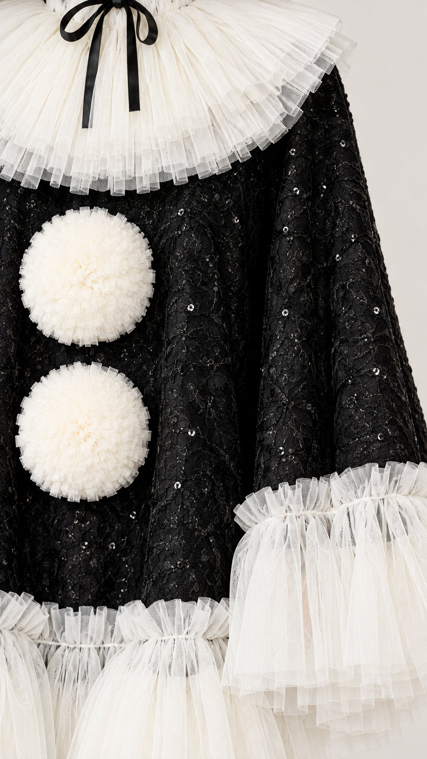 Close-up detail of textured black sequin fabric, tiered white tulle collar with black bow, and fluffy pom-pom accents