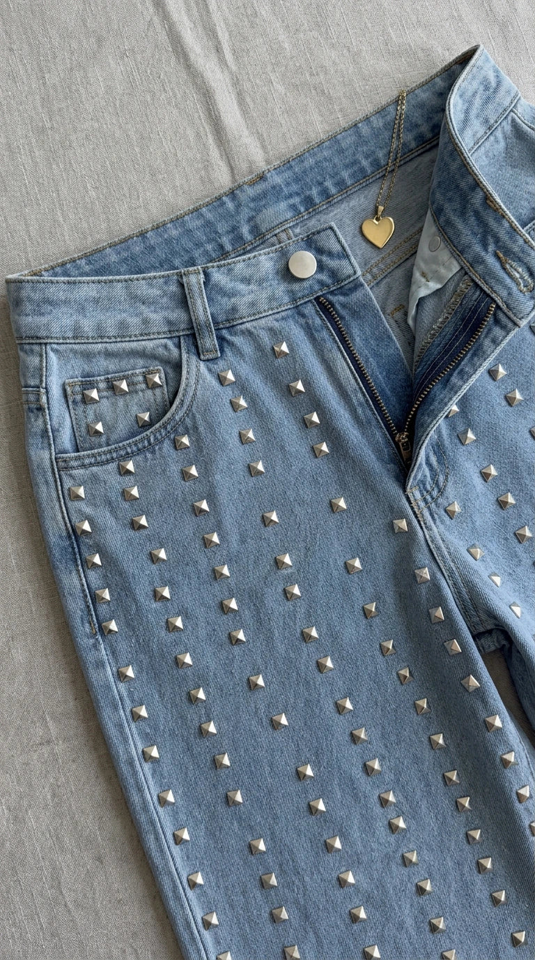 Close-up detail of light blue denim waistband, zip fly, front pocket, and silver square stud accents
