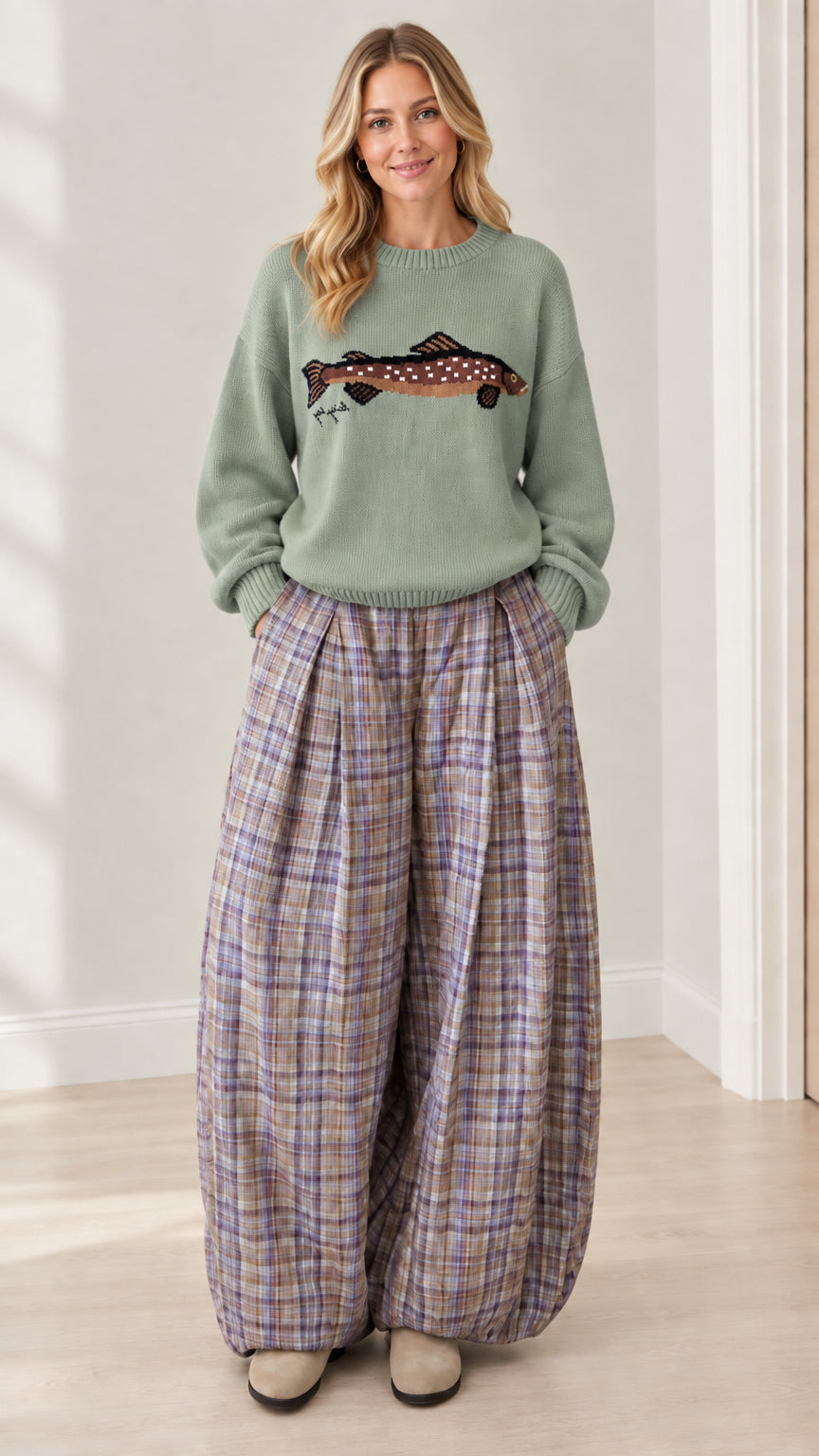 Model wearing sage green fish graphic knit sweater and pleated plaid trousers
